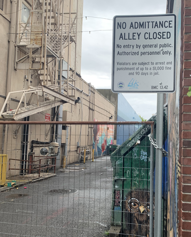 A photo of an alley in downtown Bellingham, Washington. The alley is fenced and locked with a warning against public entry.
